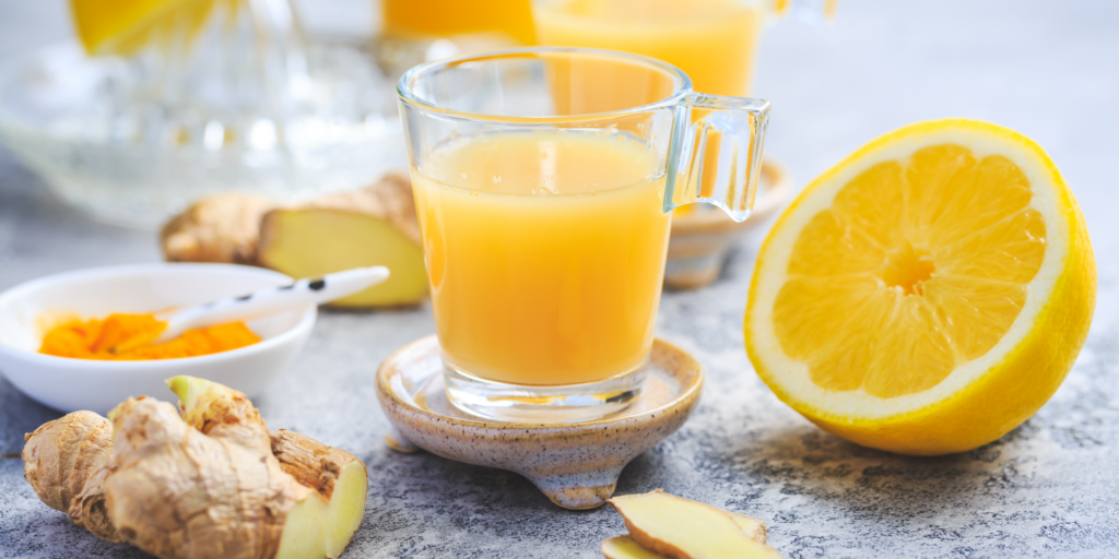 Bild, das eine Ingwerknolle und eine aufgeschnittene Zitrone zeigt. Daneben steht en kleines Glas mit einem Ingwer-Shot und im Hintergrund sieht man noch weitere Gläser mit Ingwer-Shots. Auch ein kleiner Teller mit einem orangenen Pulver ist zu sehen.