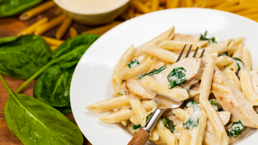 Man sieht einen Teller mit Spinat-Cashewcreme Penne. Drum herum liegt dekorative Spinatblätter.