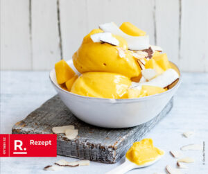 Auf dem Bild sieht man eine weiße Schüssel, gefüllt mit Mango-Kokos-Eis und mit Kokoschips garniert.